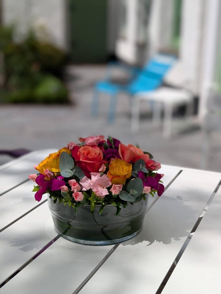 Laag rond bloemstuk met rozen, anjers en violier in een glazen schaal op een witte tuintafel
