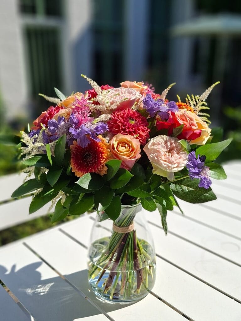 Rond en compact boeket met oranje rozen, rode dahlia's en paarse scabiosa in een glazen vaas op een witte tuintafel