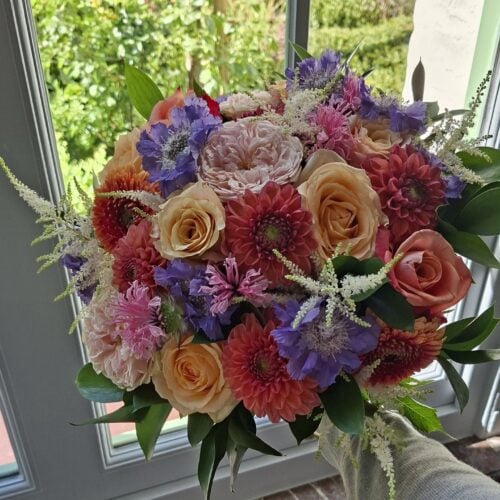 Rond handgebonden boeket met perzikkleurige rozen, koraalrode dahlia's en paarse scabiosa, bij een raam met tuinzicht