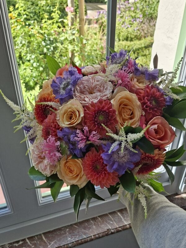 Rond handgebonden boeket met perzikkleurige rozen, koraalrode dahlia's en paarse scabiosa, bij een raam met tuinzicht