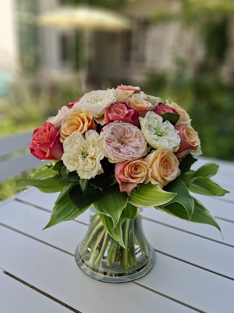 Handgebonden rozenboeket in pasteltinten in een glazen vaas op een witte tuintafel