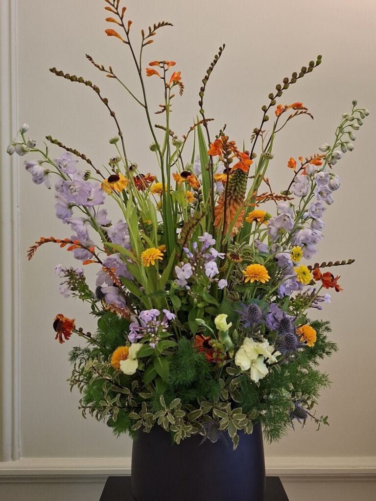 Hoog en luchtig bloemstuk met lila ridderspoor, oranje montbretia en gele zonnekruid in een donkerblauwe pot.