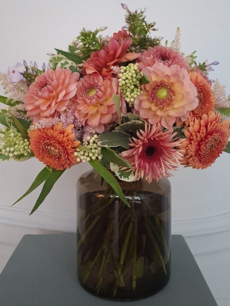Boeket met zalmroze dahlia's, gerbera's en witte astilbe in een bruine glazen vaas.