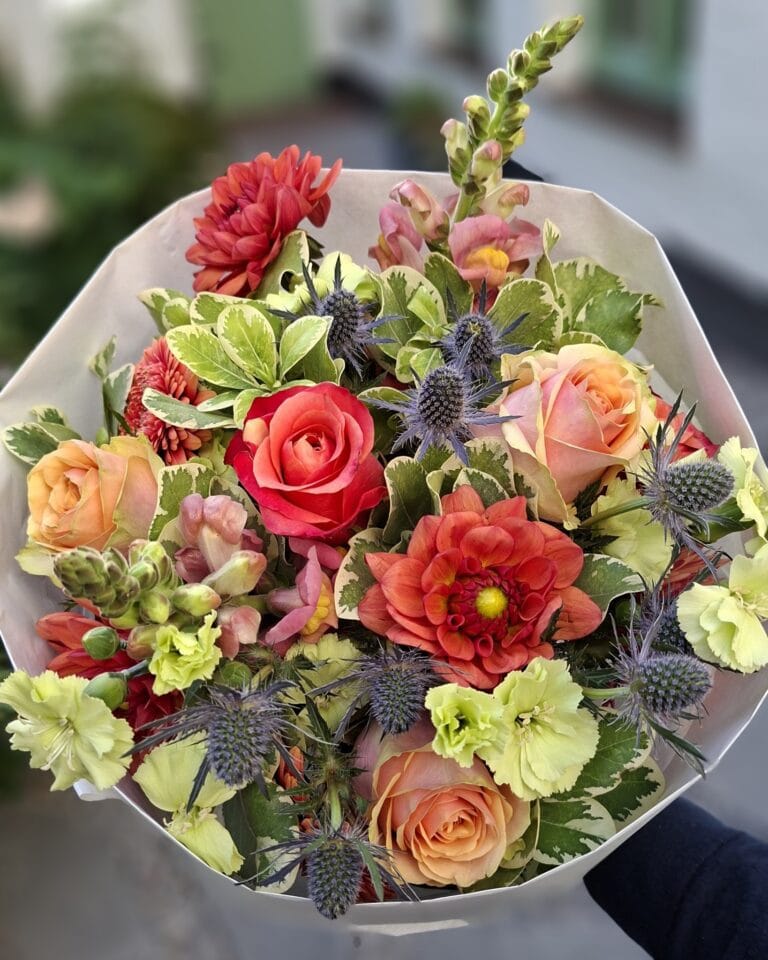 Handgebonden boeket in rood, oranje en limoengroen met rozen, dahlia, leeuwenbek en blauwe distel, in wit papier.