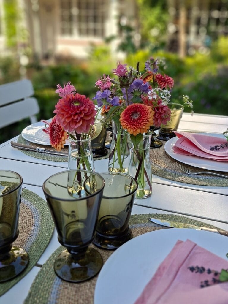 Gedekte tafel in de tuin met dahlia's in glazen vaasjes, een gedekt bord met roze servet en gerookte glazen