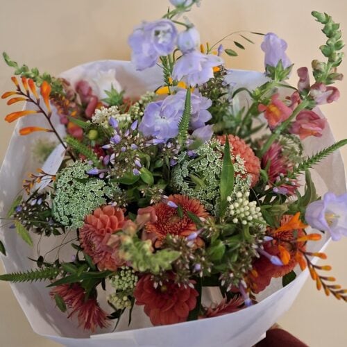 Handgebonden veldboeket met lila klokjes, oranje montbretia, wilde peen en rode dahlia's, in wit papier.