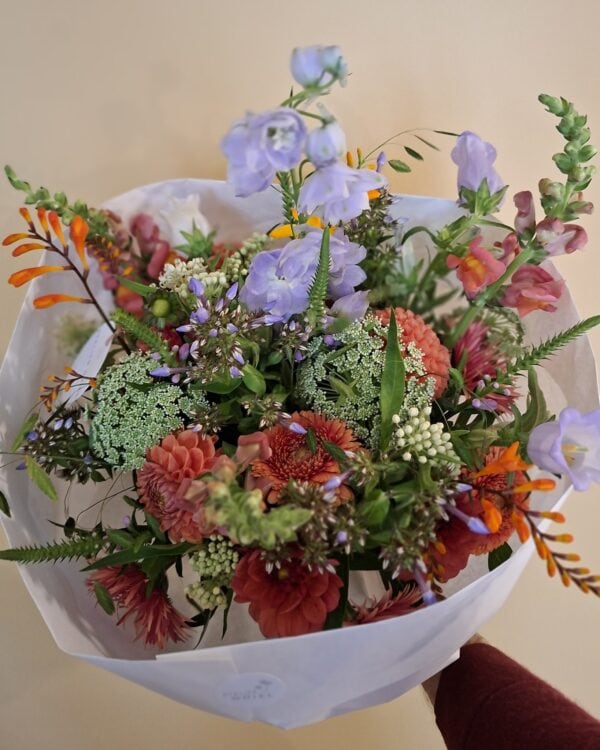 Handgebonden veldboeket met lila klokjes, oranje montbretia, wilde peen en rode dahlia's, in wit papier.