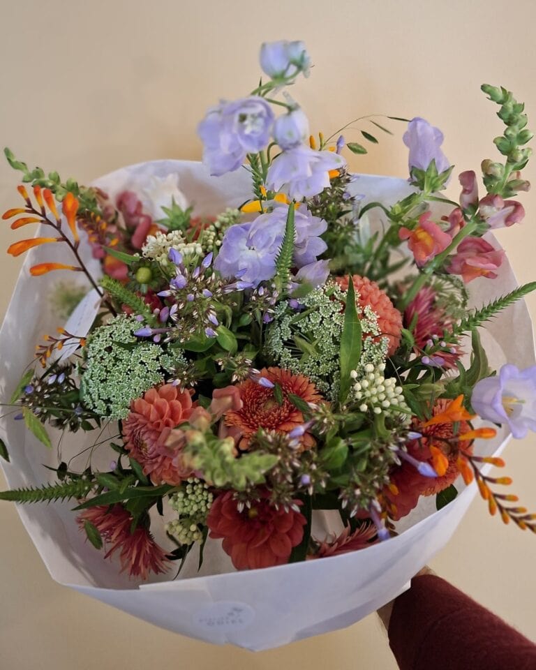 Handgebonden veldboeket met lila klokjes, oranje montbretia, wilde peen en rode dahlia's, in wit papier.